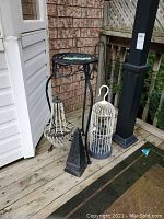 Photo showing entire group of items: black metal plant stand, hanging beaded chandelier, white birdcage candle holder, and black pyramid candle holder on wooden floor outside a house.