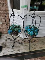 Two black metal heart-shaped hanging plant stands with wicker baskets holding blue-green faux plants, placed on wood deck against white siding.