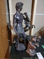 Photo of three figurative statues including standing male, rearing horse, and thinking male on table against wooden cabinet.