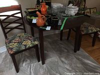 Photo showing the dark wooden rectangular table surrounded by two cushioned chairs with patterned upholstery and various glassware and pottery on the table.