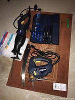 Photo showing the Mastercraft reciprocating saw with attached blade, the electric skill saw, a blue soft carrying case with multiple blades, and a separate loose blade laid on a carpet surface.