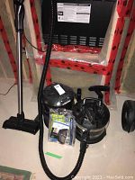 Photo showing two main units: a cylindrical vacuum cleaner and an air cleaner with control panel top, along with vacuum hose and attachments on a concrete floor near basement insulation.