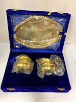 Boxed brass cream and sugar set with a matching tray, all wrapped in plastic inside a blue velvet-lined box.