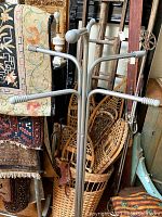 Metal coat tree with six legs base and six arms for hanging coats, shows ribbed detail on arm ends and base feet, standing on wooden floor near wicker basket and other household items.