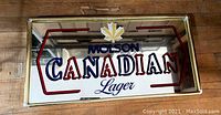 Photo of the vintage Molson Canadian Lager beer framed mirror showing the brand logo with maple leaf and the colored lettering on the mirror surface, framed in gold-tone metal.