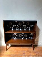 Front view of the vintage mid century modern wooden bookcase showing two shelves and Marimekko floral wallpaper backing in black and grey on white.