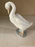 Glossy white porcelain goose figurine with peach beak and feet standing on gray base under neutral lighting