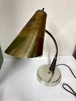 Desk lamp showing brass cone-shaped shade, curved flexible neck, and round metal base.