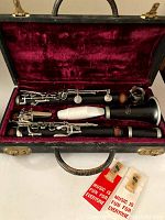 Clarinet in three parts inside open black hard case with red velvet lining, includes two reeds in packaging.