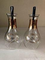 Vintage Pyrex glass oil and vinegar containers with gold band decoration and black stoppers, shown side by side on a neutral surface.
