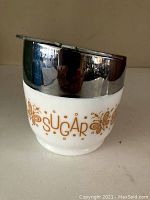 Side view of vintage sugar bowl showing white ceramic base with gold floral decoration and text 'SUGAR' plus reflective metal lid on top.
