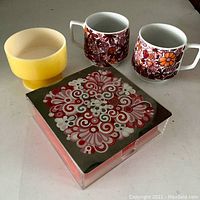 Photo showing all items in the lot: two floral ceramic mugs, yellow bowl, and square metal napkin box with retro design.