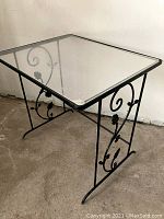 Photo of a square vintage side table with a black decorative metal base and clear plexiglass top on a concrete floor against a white wall.