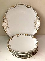 Stack of six antique white plates with scalloped and embossed edges featuring gold accents.