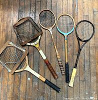 Five vintage wooden tennis racquets laid on wooden floor. Two racquets have wooden presses clamped on the heads with metal hardware. Grips vary in color and material, showing signs of use.