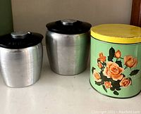 Three vintage metal canisters: two brushed aluminum with black lids, one decorated green canister with yellow lid featuring orange roses.
