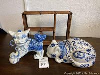 Photo showing porcelain Foo dog statue with blue and white intricate design alongside sleeping cat statuette and bamboo-style shelf in background.