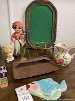 Wide shot showing the entire assortment of items on a table, including the pitcher, head vases, metal dustpan, fish-shaped dish, frame, stand, matchboxes.