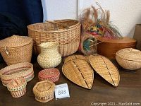 Photo showing assortment of woven baskets varying in size and style including leaf-shaped trays, small round and hexagonal baskets, and a larger two-handled basket with woven mask and bentwood box in background