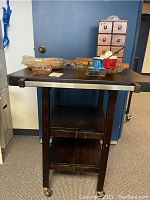 Front view of the dark brown butcher block kitchen island on casters, displaying the foldable side and bottom shelf. Various kitchen items placed on top.