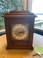 Wooden mantel clock with ornate metal clock face and brass handle on top, showing full clock face and wooden body.