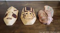Three masks displayed on wooden surface: two wooden masks with painted indigenous designs and one earthenware half skull half face mask with pastel colors