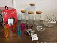 Full view of auction lot including electric kettle box, four textured glass canisters with wooden lids, small glass canister, four colored glass tumblers, smoked purple ramekin, fishtail glass serving dish, small metal dish with lid.