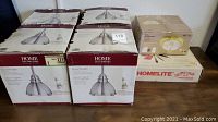 View of five boxed light fixtures: four Home Decorators Collection dome pendants and one Hampton Bay flush mount fixture, alongside a boxed Homelite Woodcutter's Kit, displayed on a table.