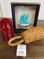Photo showing the red glass shade, framed blown glass ship with blue sails inside a black frame, and heavy jute buoy lying on a wooden surface.