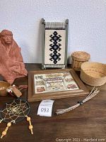 Full view of lot items arranged on flat surface showing terra-cotta statue, baskets, dreamcatcher, framed piece, small bowl, tapestry and carved wooden tool.
