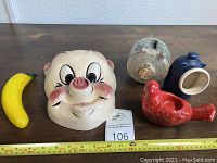 Wide view showing Porky Pig mask, speckled piggy bank, blue salt pig, red bird tealight holder, and yellow glass banana.