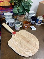 Pizza peel and assorted bowls, mugs, and plant displayed on table.