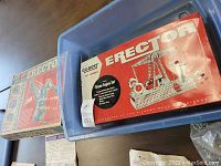 Two vintage erector set boxes stacked inside a plastic container, primarily red and blue colored boxes with mechanical model illustrations.