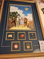 Full view of framed lithograph with characters Dorothy, Scarecrow, Tin Man and Lion walking on Yellow Brick Road and six pins below including ruby slippers and heart-shaped clock.