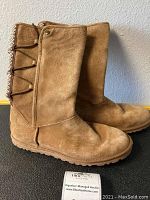 Side profile of pair of brown suede tall UGG boots with visible lace-up and decorative button on the outer side. Shows the overall shape and wear level including creased toe area.