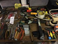 Overview of assorted tools including locking pliers, staple gun, hammers, measuring tapes, clamps, and rolls of tape on a wooden table.