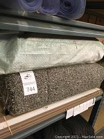 Rolled carpet remnants in beige and gray colors with rough cut edges, placed on shelf with rolled plastic carpet runners and moving blankets above.