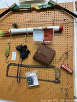 Image showing Martin Archery Cougar compound bow with green and yellow color and logo on handle, hanging on pegboard along with accessories including binoculars, quiver tube, leather bag, hand saw, styrofoam cutter