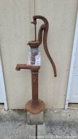 Tall rusted metal pump featuring a large curved handle and spout, mounted on a round base, showing heavy rust.