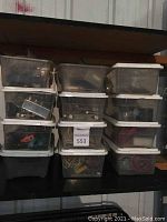 Three stacks of smaller clear plastic bins with white lids. Bins contain assorted hardware, metal parts, and electrical components. Visible items include screws, bolts, hoses, wires, and metal brackets.