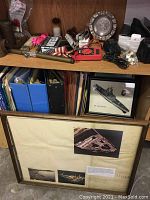 Wide view showing multiple small collectibles including brass tubes, papers, framed photos and artwork, and various small tools on wooden shelf