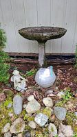Photo of concrete birdbath with reading fairy statue and globe on metal stand surrounded by rocks on the ground.