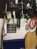 Wall-mounted vintage wooden-handled scythe with metal blade, hanging near a black pegboard with various household items including hangers, cap, and cables.