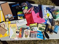 Lot of assorted crafting, baking, and holiday decor items arranged on white table outdoors. Includes colored paper, greeting cards, cork board, sewing thread, tape, and decorations.