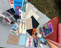 Wide view of the crafting lot including papers, color powders, glue sticks, tags, and clear acrylic items on a table outdoors.