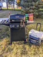 Full view of Dyna-Glo gas grill with 3 burners, smoker box holder, grilling utensils on side shelf, three packs of Charcoal Companion smoking chips on top, and MaxCold rolling cooler on ground nearby.