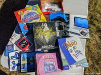 Lot contents including Cranium, The Newlywed Game, Taboo, Settlers of Catan Card Game boxes, On The Dot card game, multiple fidget spinners, laptop swivel stand, and books in a pile on the ground outdoors.