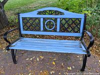 Full front view of iron and wood outdoor bench set on paved area outside. Shows curved iron arms, light blue wood seat slats and decorative backrest with lattice and circular iron designs.