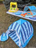 Photo showing the blue and white striped portable pop-up baby beach tent set up outdoors with its carry bag visible on the ground beside it, and three inflatable baby floats on a folding table behind the tent.