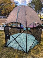 Fully assembled hexagonal pop-up playard with domed sun shade outdoors on grass showing mesh sides and zipped canopy.